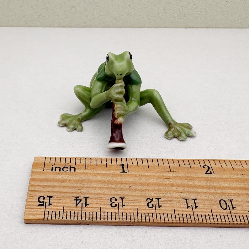 May include: A small, green ceramic frog figurine playing a brown and white instrument. The frog is posed with its legs spread and is approximately 1 inch in length. The figurine is on a white surface with a wooden ruler in the foreground.