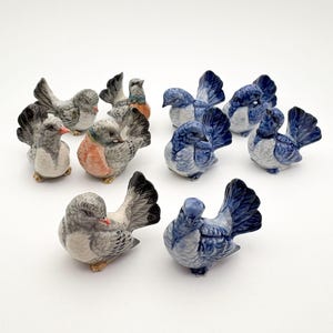 Może przedstawiać: Kolekcja ceramicznych figurek ptaków. Ptaki są w różnych pozach, niektóre zwrócone przodem, a inne bokiem. Ptaki są pomalowane w odcieniach niebieskiego, szarego i białego, niektóre mają pomarańczowe akcenty.