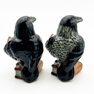 May include: Two black raven figurines reading books. The raven on the left has black feathers, while the raven on the right has gray feathers. Both ravens are perched on open books with brown covers.