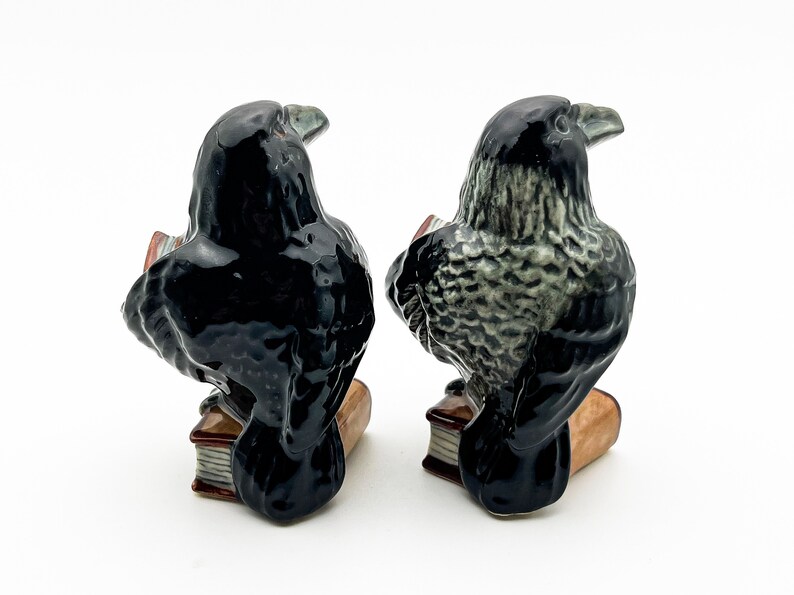 May include: Two black raven figurines reading books. The raven on the left has black feathers, while the raven on the right has gray feathers. Both ravens are perched on open books with brown covers.