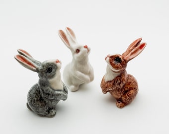 May include: Three ceramic rabbit figurines. One is white, one is gray, and one is brown. The rabbits are all in a sitting position with their ears up.