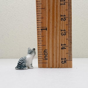 May include: A small, ceramic cat figurine in a seated position, with gray and white stripes. The figurine is approximately 1 inch tall, next to a wooden ruler.