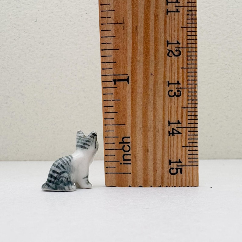 May include: A small, ceramic cat figurine in a seated position, with gray and white stripes. The figurine is approximately 1 inch tall, next to a wooden ruler.