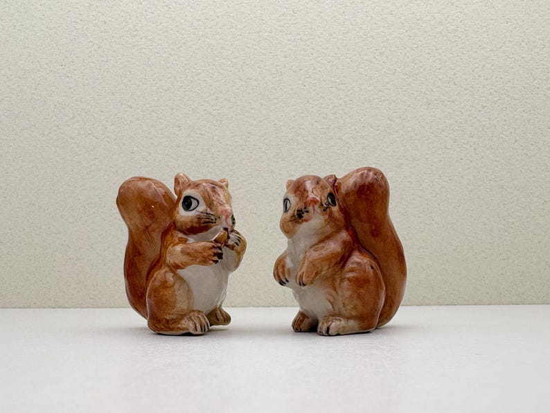 Peut inclure: Deux figurines de l'&eacute;cureuil en c&eacute;ramique. Un &eacute;cureuil tient une noix. Les &eacute;cureuils sont bruns et blancs avec des d&eacute;tails. Ils ont de grandes queues touffues et sont pos&eacute;s sur une surface blanche.