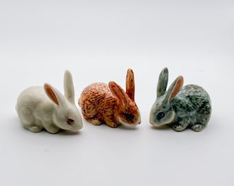 Könnte beinhalten: Drei Keramik-Hasenfiguren, eine weiße, eine braune und eine graue, sind in einer Reihe auf einer weißen Oberfläche angeordnet.