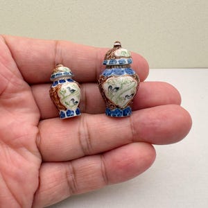 Op de afbeelding: Twee miniatuur decoratieve keramische vazen met deksels. De vazen zijn bruin, blauw en wit, met een vogel- en bladerenontwerp. De kleine vazen zijn waarschijnlijk voor display of decoratieve doeleinden.