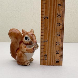 Peut inclure: Figurine de petit &eacute;cureuil en c&eacute;ramique, principalement marron avec un ventre blanc, tenant une noix. La figurine mesure environ 5 cm de haut. Une r&egrave;gle en bois avec des mesures en pouces est &agrave; c&ocirc;t&eacute; de l'&eacute;cureuil.