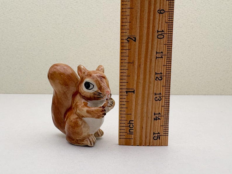 Peut inclure: Figurine de petit &eacute;cureuil en c&eacute;ramique, principalement marron avec un ventre blanc, tenant une noix. La figurine mesure environ 5 cm de haut. Une r&egrave;gle en bois avec des mesures en pouces est &agrave; c&ocirc;t&eacute; de l'&eacute;cureuil.