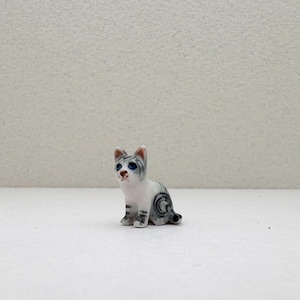 May include: A small, ceramic cat figurine in a seated position. The cat is primarily white with gray tabby markings and blue eyes. The figurine has a detailed, hand-painted appearance, with a small, black tail.