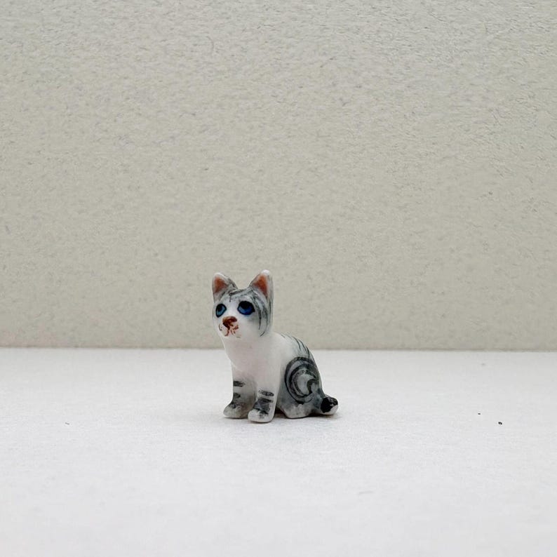 May include: A small, ceramic cat figurine in a seated position. The cat is primarily white with gray tabby markings and blue eyes. The figurine has a detailed, hand-painted appearance, with a small, black tail.