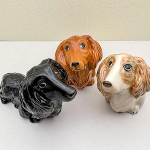 Puede incluir: Tres figuritas de dachshund de cerámica. Una es negra, otra es marrón y otra es blanca con marcas marrones.