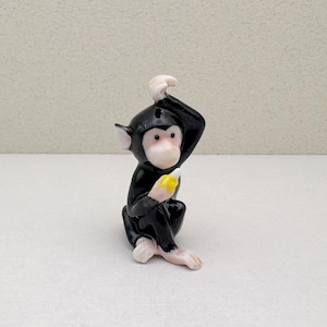 May include: A black porcelain figurine of a monkey sitting with its hand raised to its head, holding a yellow banana. The monkey has a white face and pink hands and feet.