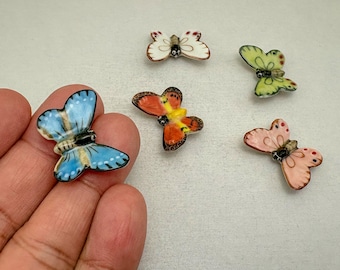 Peut inclure: Cinq petites figurines de papillons en céramique de différentes couleurs, notamment bleu, blanc, orange, jaune, vert et rose. Chaque papillon présente des ailes peintes détaillées avec des contours noirs et un corps central.