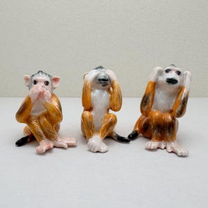 Peut inclure: Trois figurines de singes en céramique en position assise. Le premier singe se couvre la bouche, le deuxième se couvre les yeux et le troisième se couvre les oreilles. Chaque singe a un visage et un torse blancs avec des détails de fourrure orange et marron.