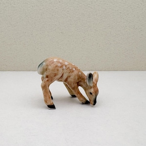 May include: A small ceramic figurine of a fawn, painted in shades of brown and white with black hooves. The fawn is depicted in a crouching position, with its head lowered as if it is grazing.