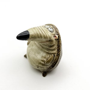May include: A ceramic figurine of a cartoon hedgehog with a brown and white striped body, black nose, and large eyes. The figurine is looking to the right and has a happy expression.