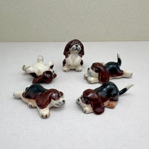 May include: A set of five small ceramic dog figurines featuring brown, black, and white markings. One dog sits upright while the other four are depicted in various lying or resting positions, all with painted facial features and floppy ears.