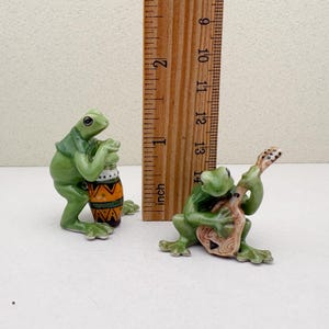 May include: Two ceramic frog figurines. One frog plays a drum with orange, yellow, and brown patterns. The other frog plays a stringed instrument. The figurines are green and measure approximately 1 inch tall.
