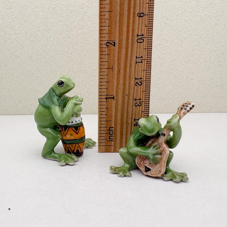 May include: Two ceramic frog figurines. One frog plays a drum with orange, yellow, and brown patterns. The other frog plays a stringed instrument. The figurines are green and measure approximately 1 inch tall.