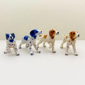 以下が含まれることがあります： 異なる色のパターンを持つ、4つの犬の磁器製置物。犬は白い表面に立っています。犬はすべて正面を向いており、頭を少し横に傾けています。犬はすべて白いベースコートに茶色、青、またはオレンジの斑点があります。