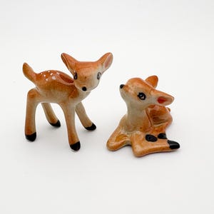Peut inclure: Deux figurines de cerfs en céramique. L'un des cerfs est debout, l'autre est couché. Le cerf debout a des taches blanches et des sabots noirs. Le cerf couché a des taches noires sur ses sabots. Les deux cerfs sont de couleur marron et beige.