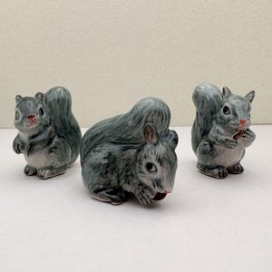 May include: Three ceramic squirrel figurines in shades of gray and white. Two squirrels are upright, and one is lying down. One squirrel holds a small brown object. The figurines have detailed features and are set against a plain background.
