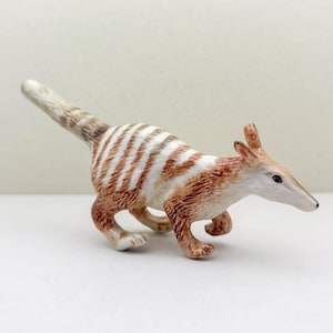 Könnte beinhalten: Detaillierte Figur eines Bilbys, eines kleinen Beuteltiers aus Australien. Das Bilby wird in einer Laufpose dargestellt, mit einem weiß-braun gestreiften Körper, langen Ohren und einer spitzen Schnauze. Die Figur steht vor einem schlichten weißen Hintergrund.