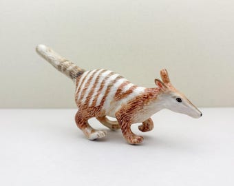 Może przedstawiać: Szczegółowa figurka bilby, małego torbacza pochodzącego z Australii. Bilby jest przedstawiony w pozie biegu, z biało-brązowym, pasiastym ciałem, długimi uszami i spiczastym pyskiem. Figurka jest ustawiona na gładkim, białym tle.