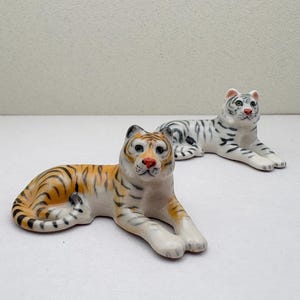 May include: Two ceramic tiger figurines. One is orange and black striped, the other is white with grey stripes. Both are in a reclining pose, with detailed facial features and realistic colouring.