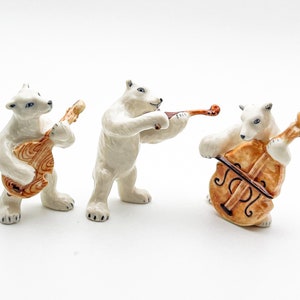 Pode incluir: Três estatuetas de cerâmica de ursos polares brancos tocando instrumentos musicais. O urso à esquerda toca guitarra, o urso do meio toca violino e o urso à direita toca violoncelo.