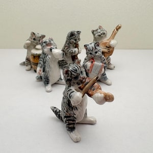 May include: A set of six small ceramic cat figurines, each painted with grey tabby stripes and white accents. The cats are depicted as musicians, with individual figures playing a violin, an accordion, a lute, a flute, and a drum, while one cat sings.