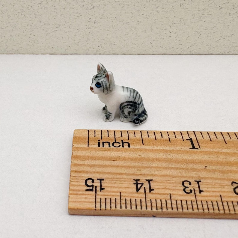 May include: A small, ceramic figurine of a seated cat. The cat is white with gray stripes and blue eyes. A wooden ruler with inch markings is in the foreground.