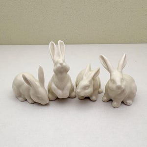 Może przedstawiać: Cztery kremowobiałe ceramiczne figurki królików w różnych pozach. Dwa króliki siedzą wyprostowane, a pozostałe dwa odpoczywają. Króliki mają długie uszy i nadają się do dekoracji wielkanocnych lub wiosennych.