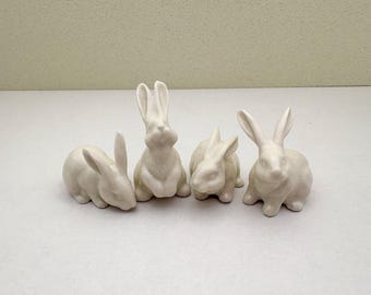 May include: Four off-white ceramic rabbit figurines in various poses. Two rabbits are sitting upright, while the other two are in a resting position. The bunnies have long ears and are suitable for Easter or spring decor.