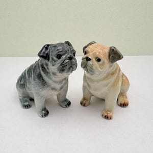 Könnte beinhalten: Zwei Bulldoggen-Figuren aus Keramik. Eine ist grau mit schwarzen Akzenten, die andere ist beige mit braunen Akzenten. Beide sitzen aufrecht mit detaillierten Gesichtern und kleinen, dunklen Pfoten. Die Figuren stehen auf einer weißen Oberfläche.