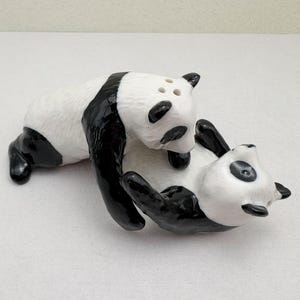 Op de afbeelding: Een zout- en peperstel van keramiek in de vorm van twee panda's. De ene panda staat over de andere heen, die op zijn rug ligt. De panda's zijn wit met zwarte vlekken rond de ogen, oren en ledematen. Drie kleine gaatjes zijn aan de bovenkant zichtbaar.