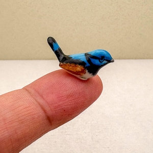 May include: A small ceramic bird figurine, mainly blue with black and orange details. The miniature bird is resting on a fingertip, highlighting its intricate design and bright colours. The figurine is a decorative item.