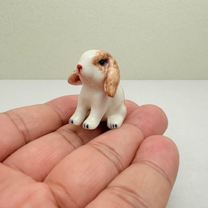 May include: A miniature ceramic figurine of a lop-eared rabbit with a white body and light brown patches on its ears and head. The rabbit has a small red nose, dark eyes, and black painted details on its paws, depicted in a seated position.