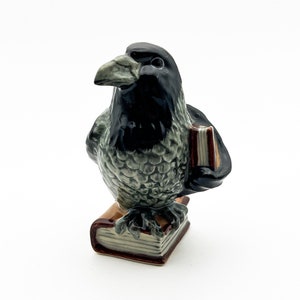May include: A ceramic figurine of a black and gray crow holding a book. The crow is perched on top of a stack of books with brown covers.