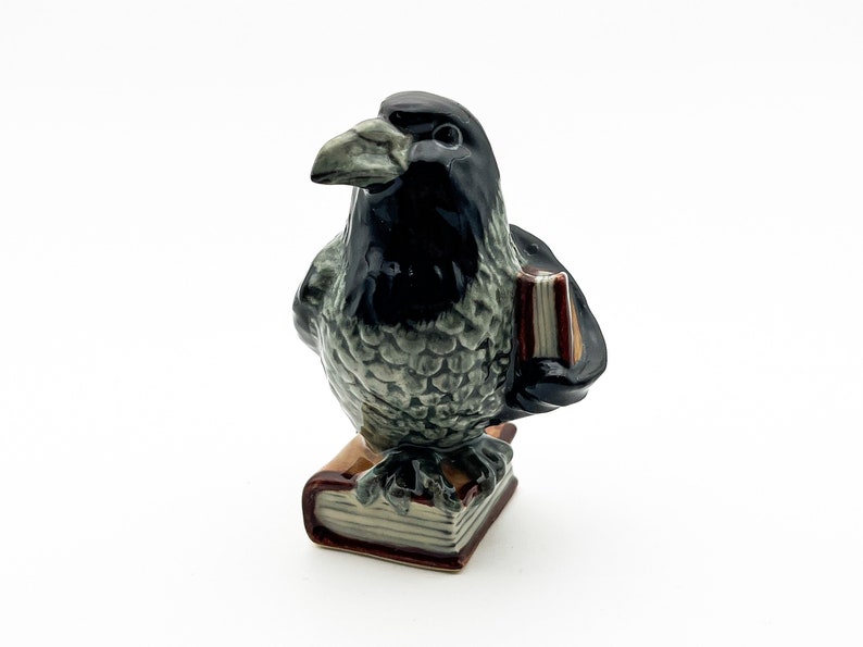 May include: A ceramic figurine of a black and gray crow holding a book. The crow is perched on top of a stack of books with brown covers.