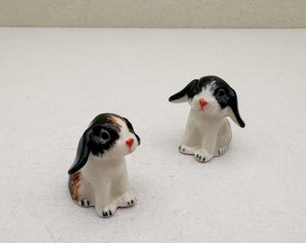 May include: Two small ceramic bunny figurines. The bunnies are white with black and brown markings on their ears and heads. They have red noses and are sitting upright. The figurines are on a white surface.