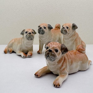 May include: Four ceramic pug dog figurines in various poses. The pugs are tan and cream-colored with black accents on their ears and faces. One pug is lying down, while the others are sitting or standing. The figurines are on a white surface.