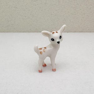 May include: A small, white ceramic deer figurine with brown spots and pink hooves. The deer has large, dark eyes and upright ears. The figurine is standing on a white surface.