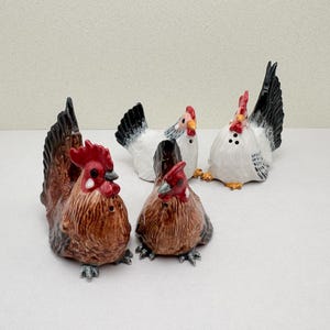 Può includere: Un set di quattro statuette di pollo in ceramica, tra cui due galli marroni con creste rosse e due galline bianche con piume della coda nere. Ogni statuetta presenta piccoli fori sulla parte superiore, indicando la loro funzione di saliera.