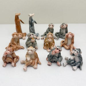 Pode incluir: Uma coleção de doze estatuetas de macaco de cerâmica em várias poses. Os macacos são pintados em tons de marrom, cinza e branco. Alguns macacos estão segurando pequenos objetos, como uma banana ou uma flor.