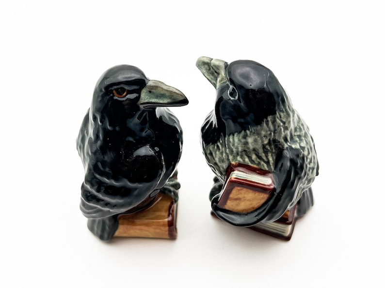 May include: Two black and gray ceramic crow figurines perched on books. The crows are facing each other and appear to be talking.