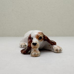 May include: A ceramic figurine of a dog, lying down with its head and paws forward. The dog has white fur with brown spots on its ears and around its eyes. The figurine has a black nose and is resting on a white surface.