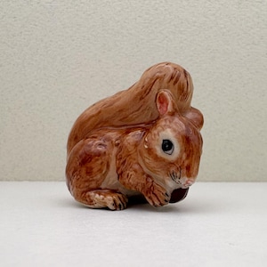 Peut inclure: Figurine en c&eacute;ramique repr&eacute;sentant un &eacute;cureuil assis. L'&eacute;cureuil a une fourrure brune avec des reflets plus clairs, une queue touffue et un petit &oelig;il sombre. Il semble tenir une noix. La figurine est sur une surface blanche.