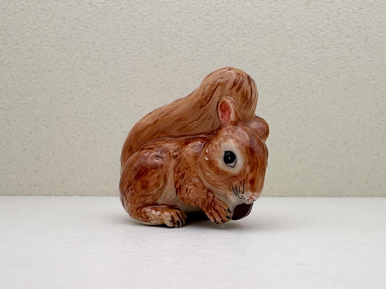 Peut inclure: Figurine en c&eacute;ramique repr&eacute;sentant un &eacute;cureuil assis. L'&eacute;cureuil a une fourrure brune avec des reflets plus clairs, une queue touffue et un petit &oelig;il sombre. Il semble tenir une noix. La figurine est sur une surface blanche.