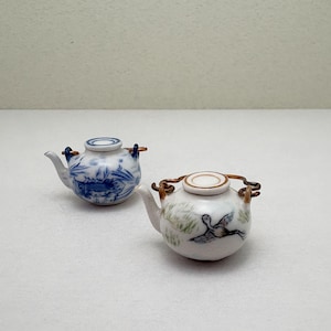 May include: Two miniature ceramic teapots. One is decorated with blue and white floral patterns, the other with a bird illustration. Both have copper-coloured handles and lids. The teapots are small and decorative.